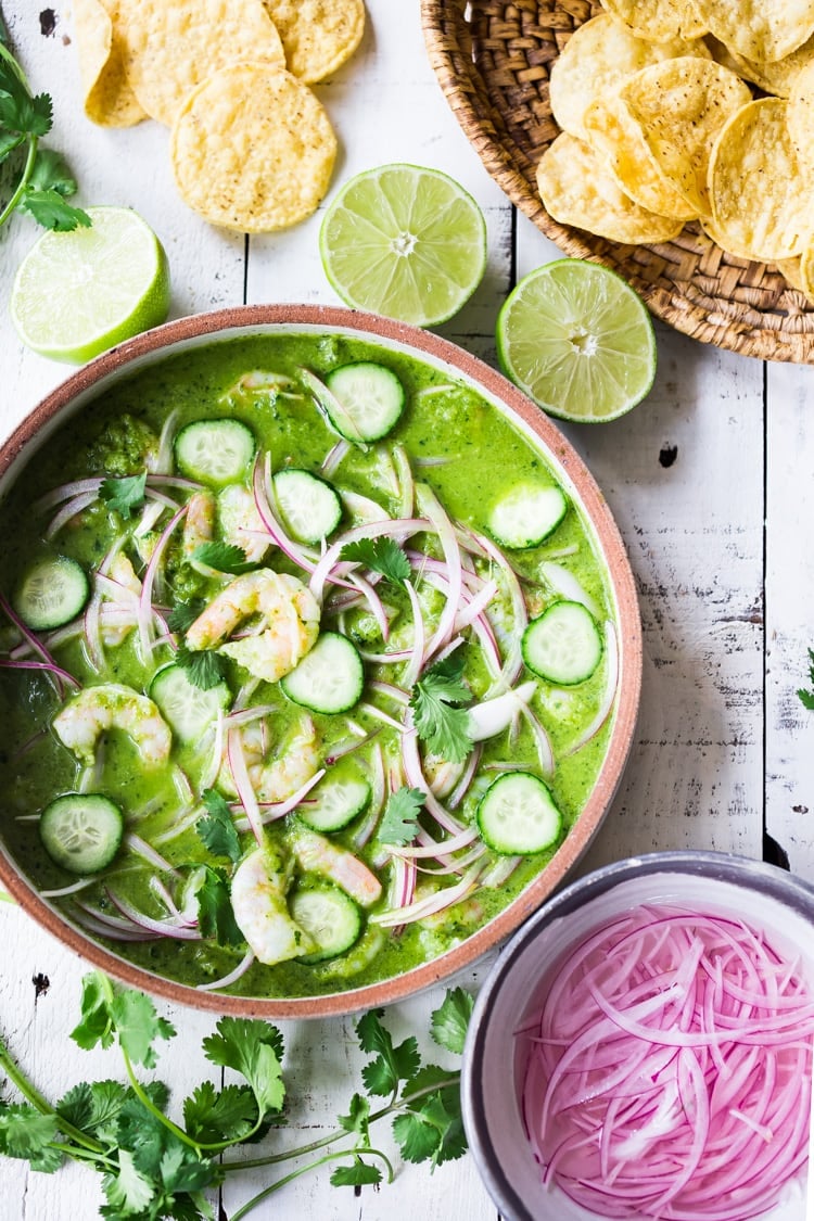 Mexican aguachile in bowl with sliced cucumber, onion, and cucumber with shrimp and cilantro next to limes and chips. 