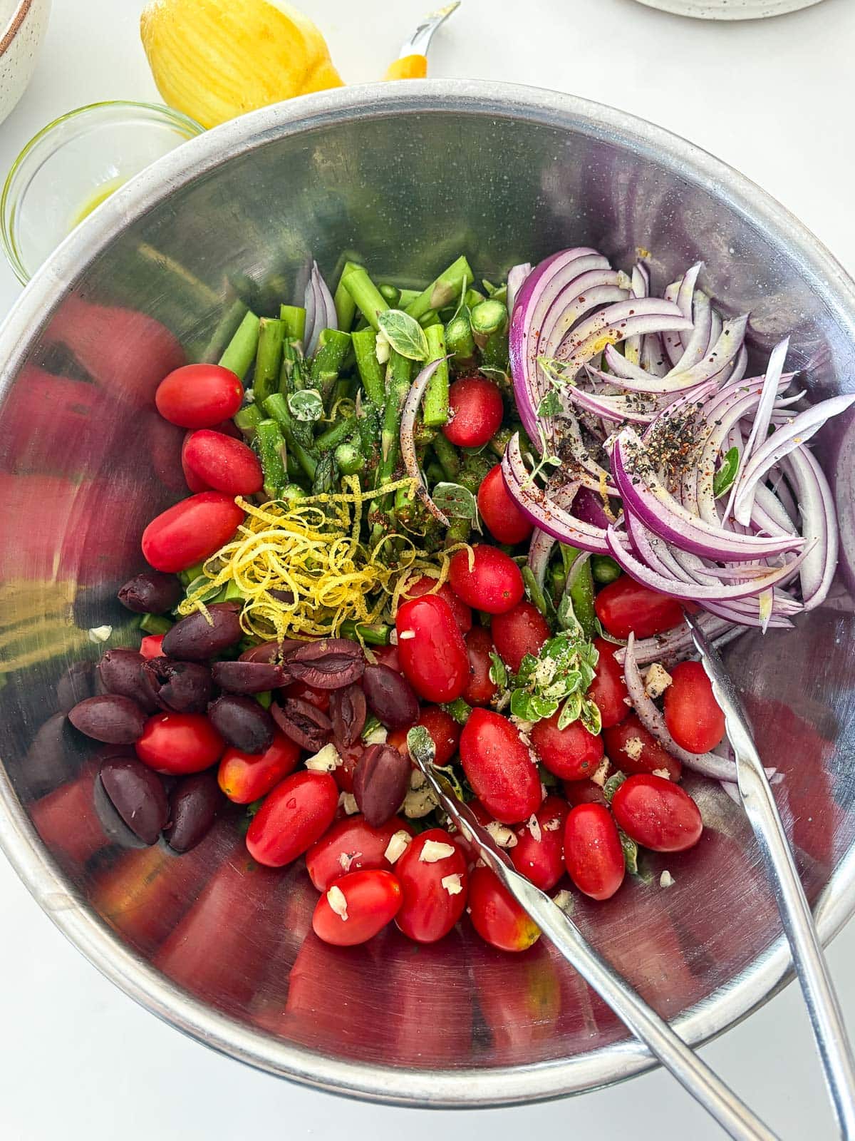 metal mixing bowl with tongs with cherry tomatoes, olives, sliced red onion, asparagus, and lemon zest. 