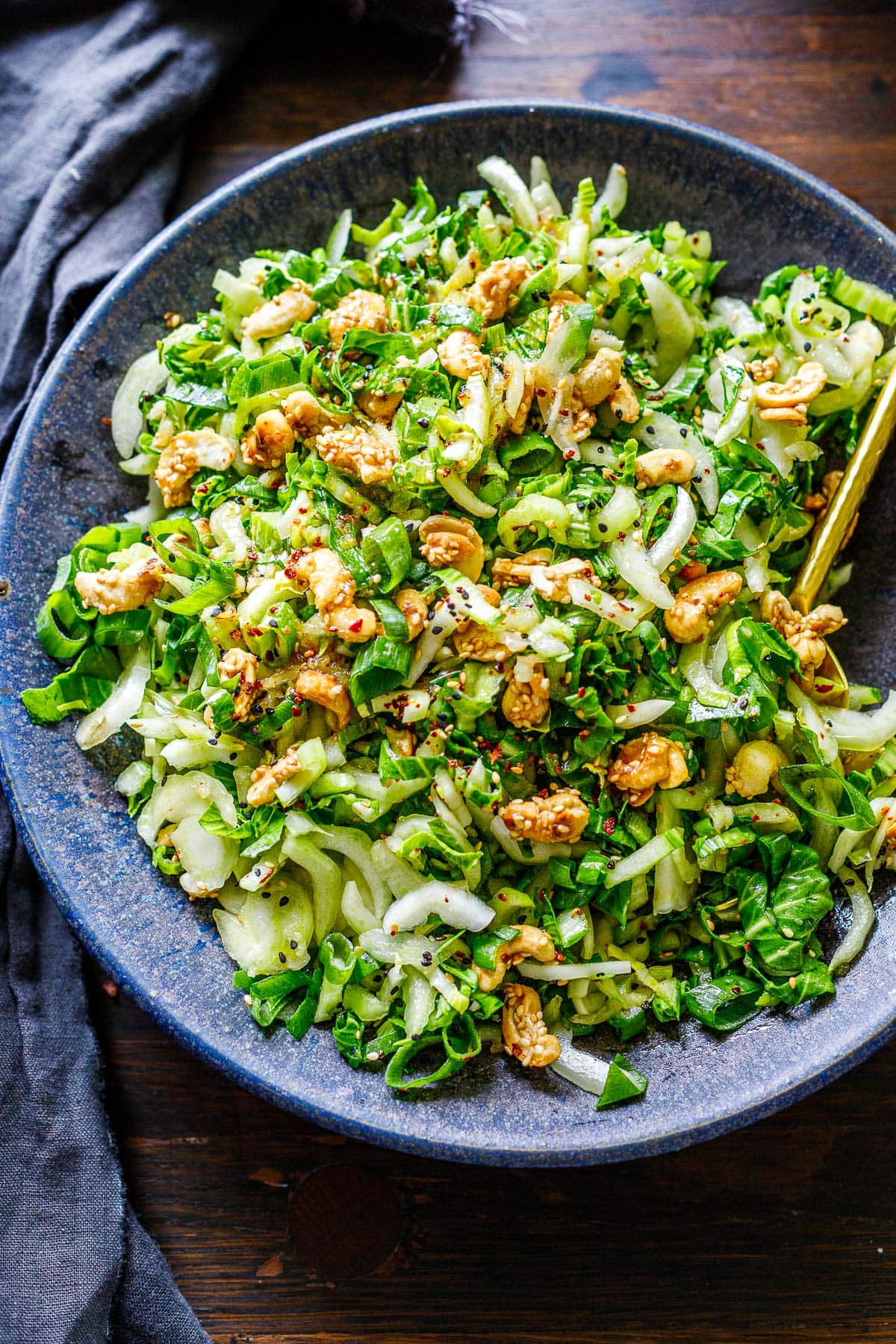 bok choy salad in a bowl with gold serving utensil, topped with sesame cashew brittle and sesame ginger dressing.
