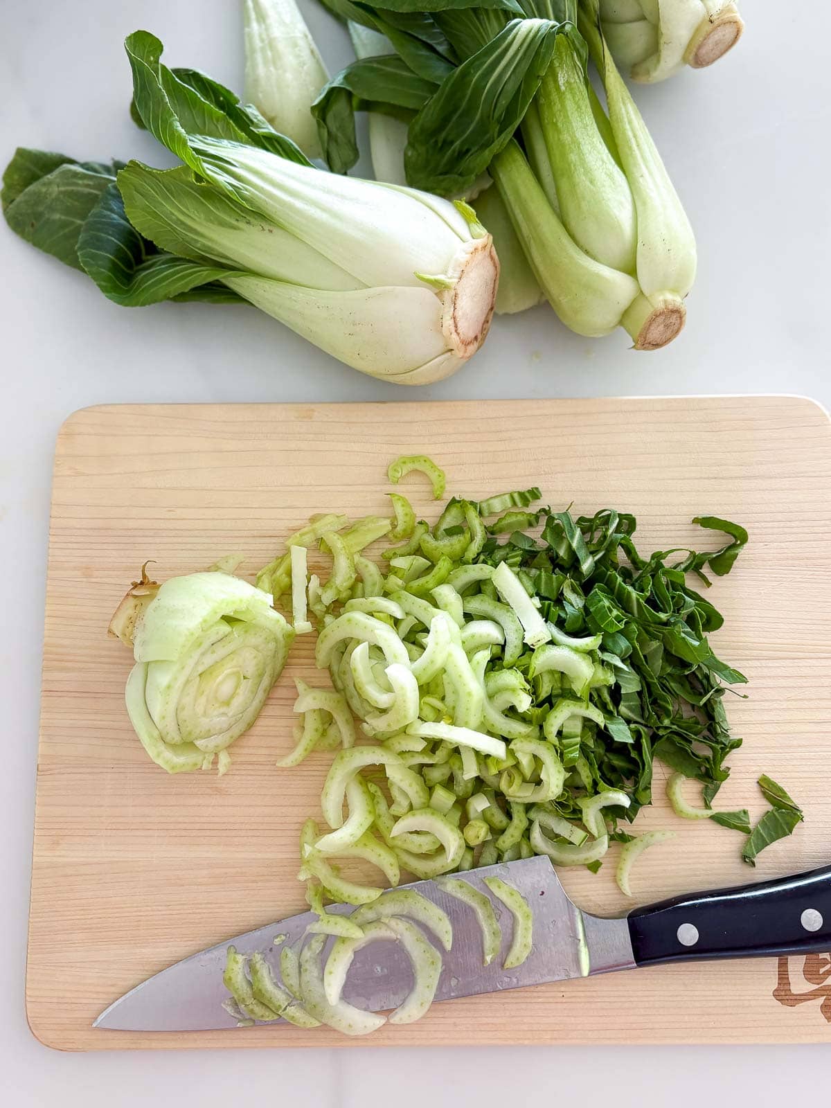 wood board and sharp knife with thinly sliced bok choy with more bok choy heads above the board.