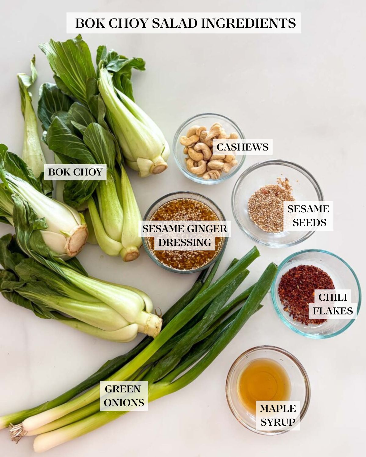 ingredients for bok choy salad labeled and neatly arranged - bok choy heads, green onions, and cashews, sesame ginger dressing, sesame seeds, chili flakes, and maple syrup in small glass bowls. 