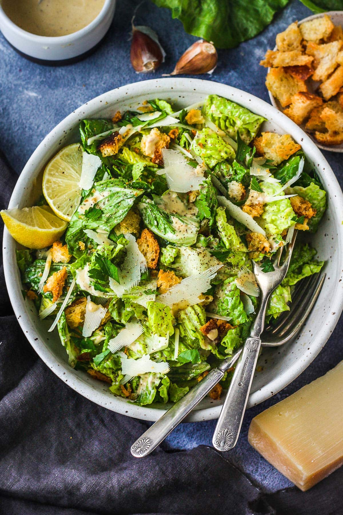 caesar salad in bowl with shaved parmesan and hand-torn croutons, lemon wedges, black pepper, caesar dressing, and forks. 