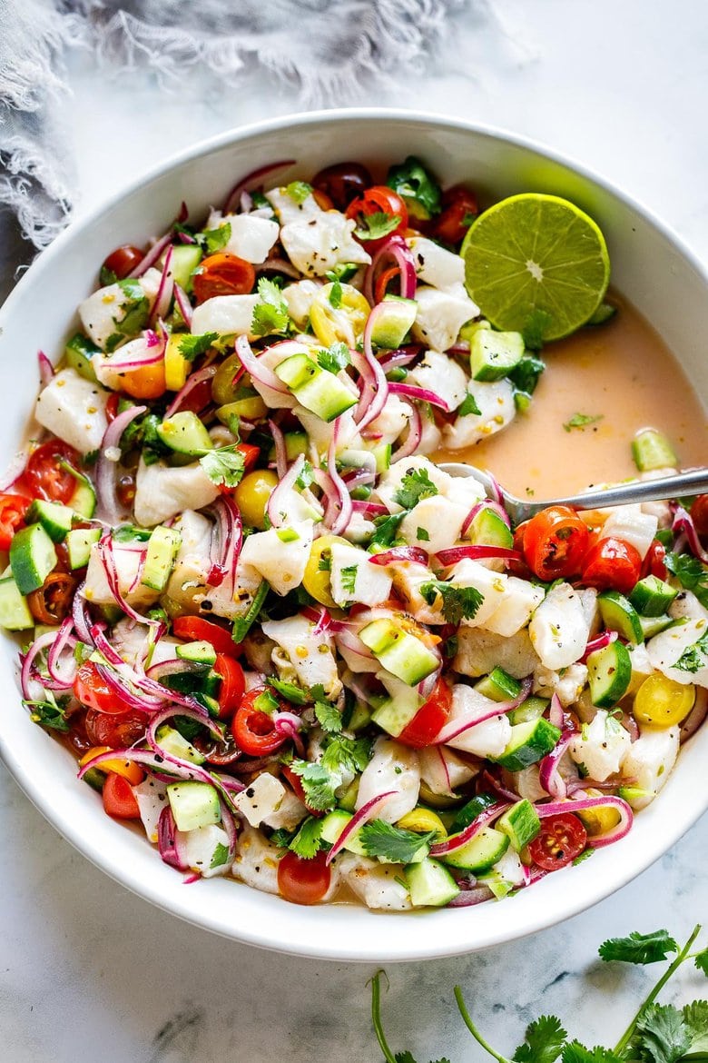 Ceviche in serving bowl with onion, tomato, cucumber, cilantro, and fresh fish with lime halve and spoon. 