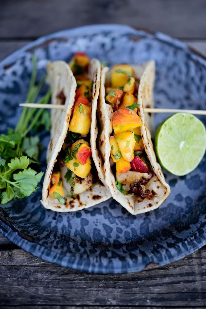 two grilled chipotle fish tacos on plate skewered together, topped with peach salsa and served with fresh cilantro and lime. 