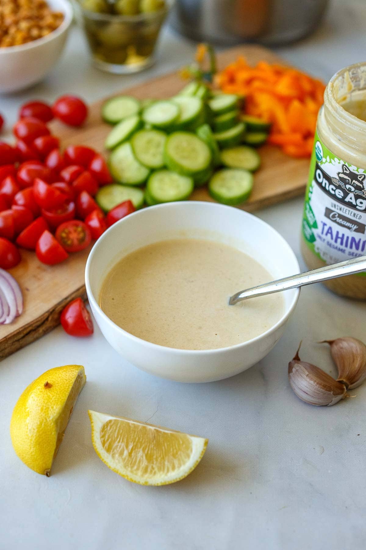 ingredients prepped on a wood board - orange bell pepper, cucumber, cherry tomatoes, and red onion, next to a jar of tahini and a small bowl of tahini sauce, lemon wedges, and garlic clove. 