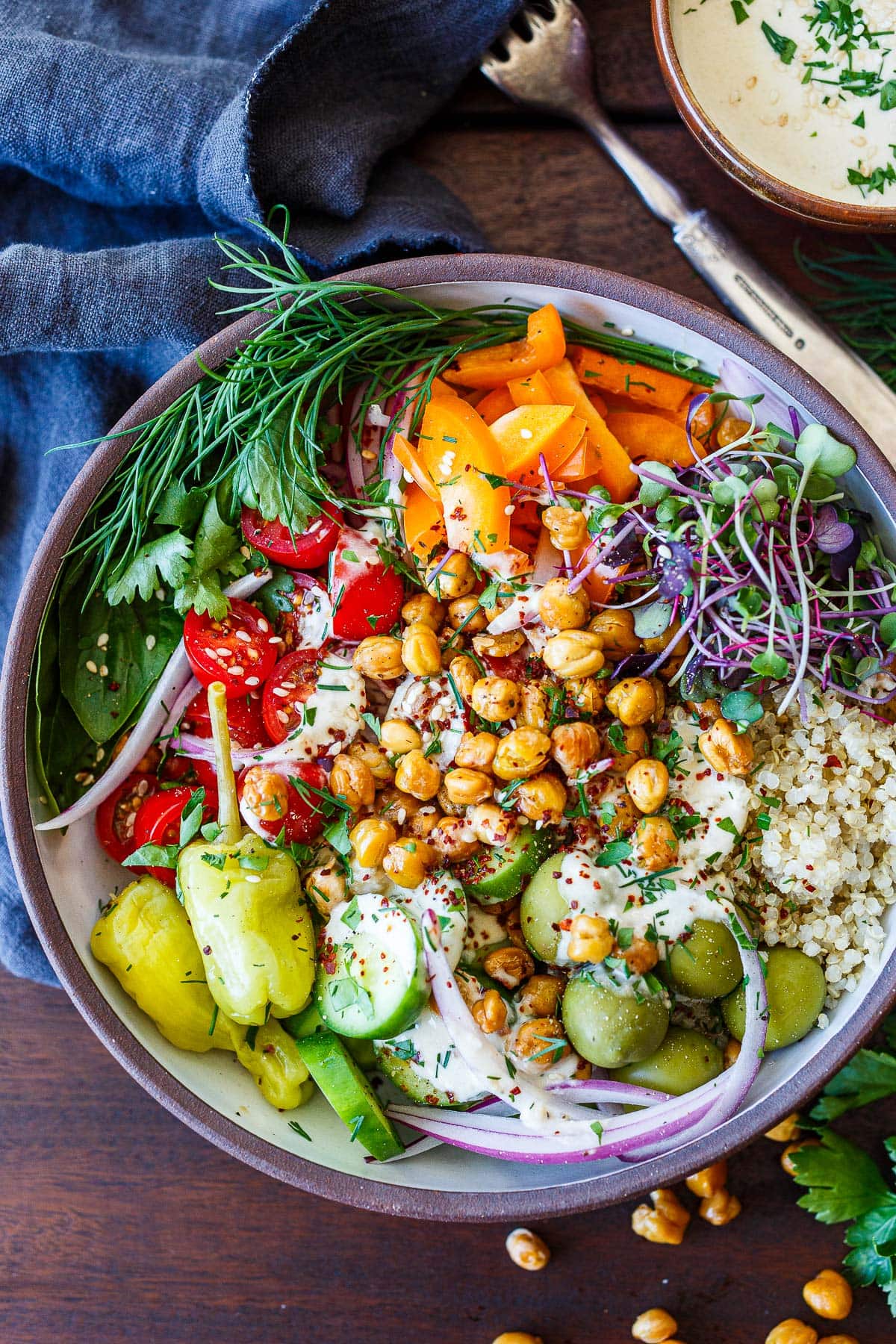 Crispy  chickpea bowl with quinoa, topped with micro greens, orange bell pepper, dill, parsley, basil, cherry tomatoes, red onion, pepperoncini, cucumber, olives, and chickpeas, with tahini sauce.