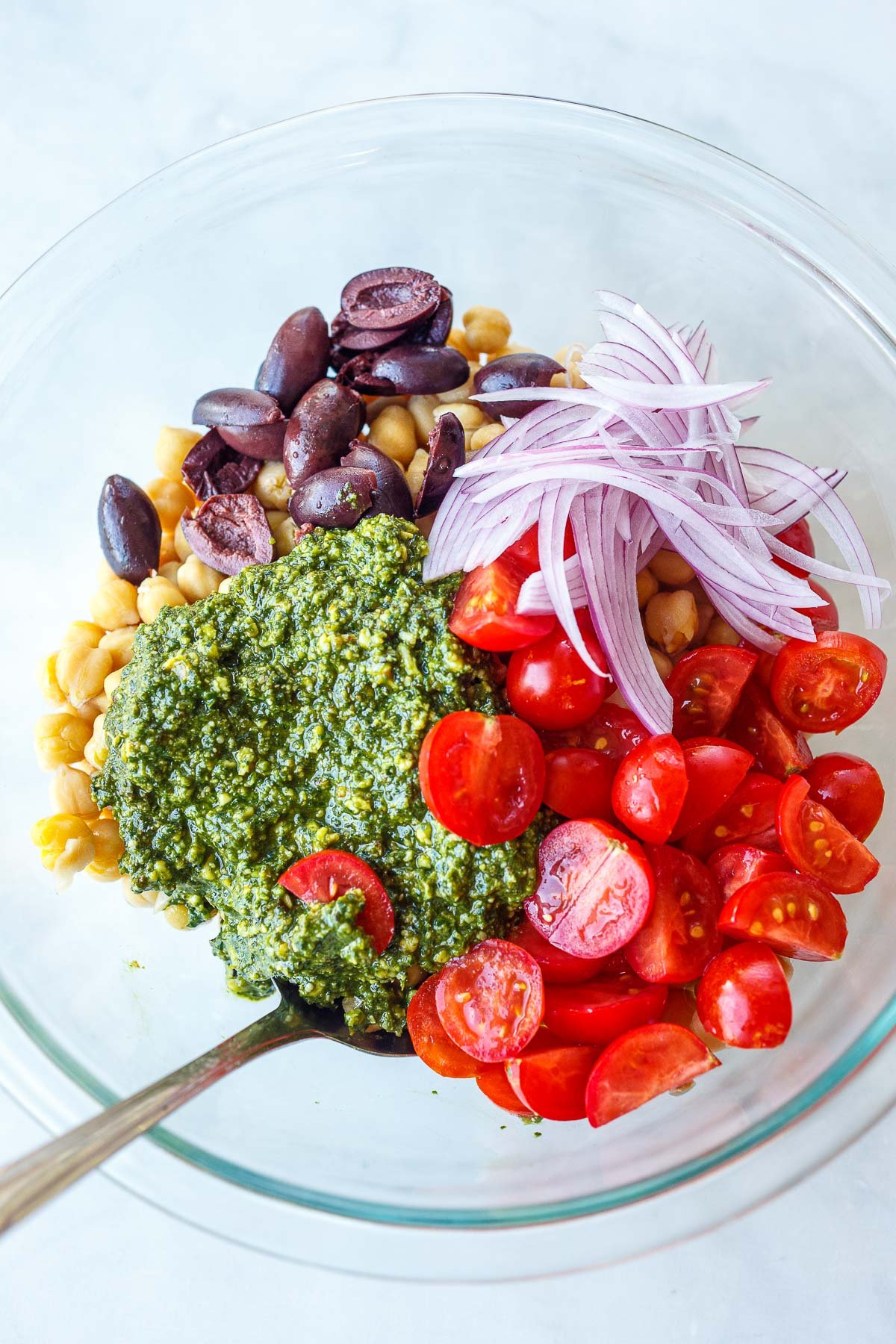 glass mixing bowl with chickpeas, olives, red onion slices, cherry tomatoes, and pesto. 