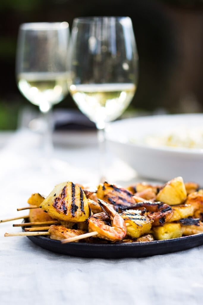 grilled shrimp skewers with pineapple stacked on plate in front of two glasses of white wine. 