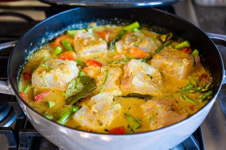 white fish added to pot of thai fish curry with colorful veggies and herbs. 
