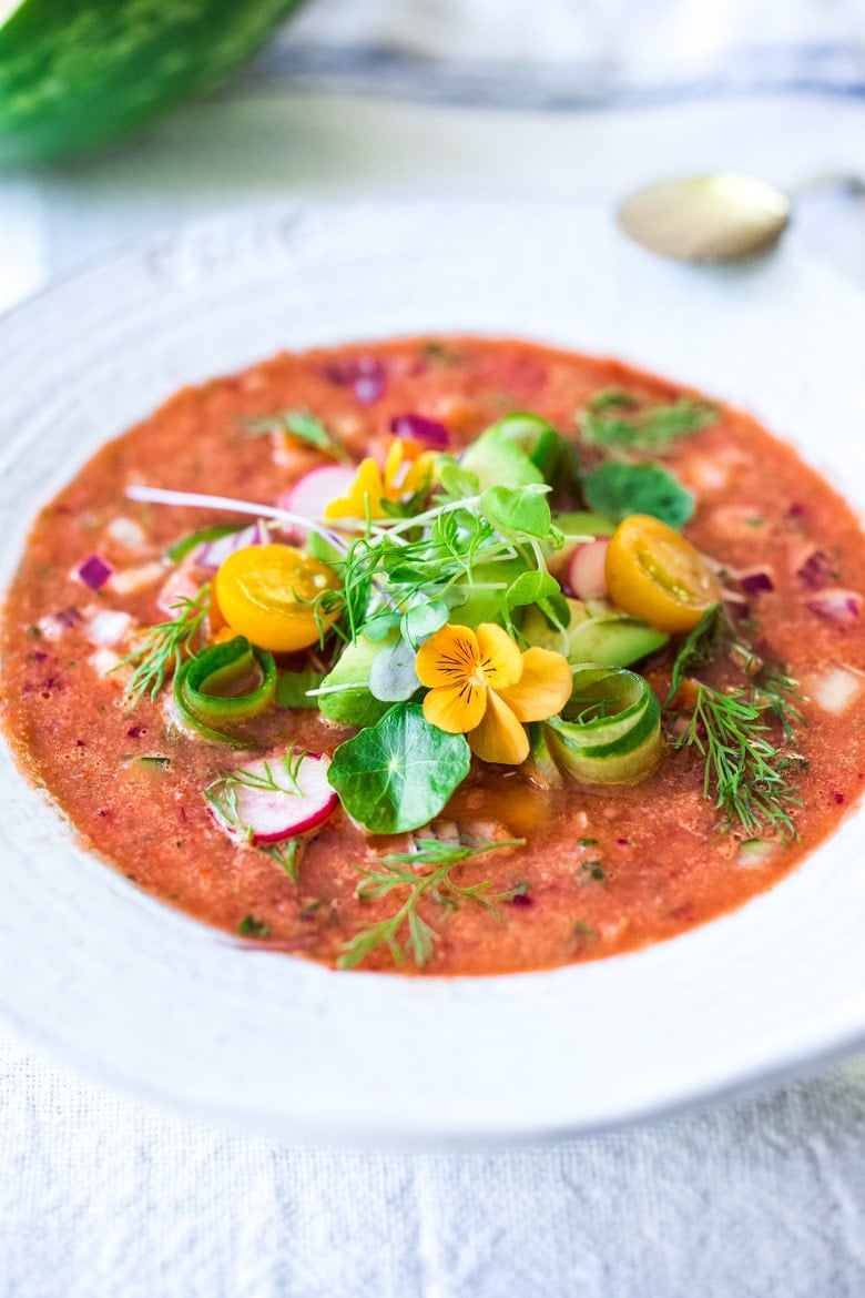 watermelon gazpacho in bowl garnished with spiralized cucumber, cherry tomatoes, fresh herbs, and yellow flowers. 